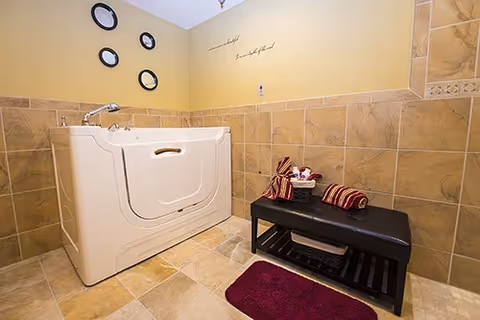 A walk-in bathtub in a tiled bathroom corner with beige and brown tiles. There is a black bench with folded towels and a basket on it, a burgundy bath mat on the floor, and four round mirrors on the wall above the bathtub. The wall is painted light yellow with a decorative quote.