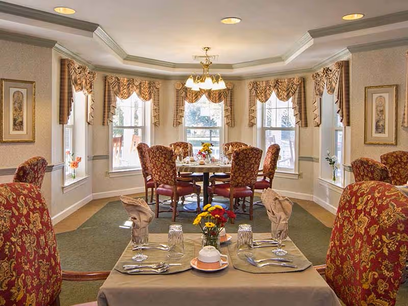 A formal dining room with a round table set for a meal, surrounded by six upholstered chairs with red and gold patterned fabric. The room has multiple windows with decorative valances, framed artwork on the walls, and a chandelier hanging from the ceiling. The foreground shows a table set with utensils, glasses, napkins, and a small flower arrangement.