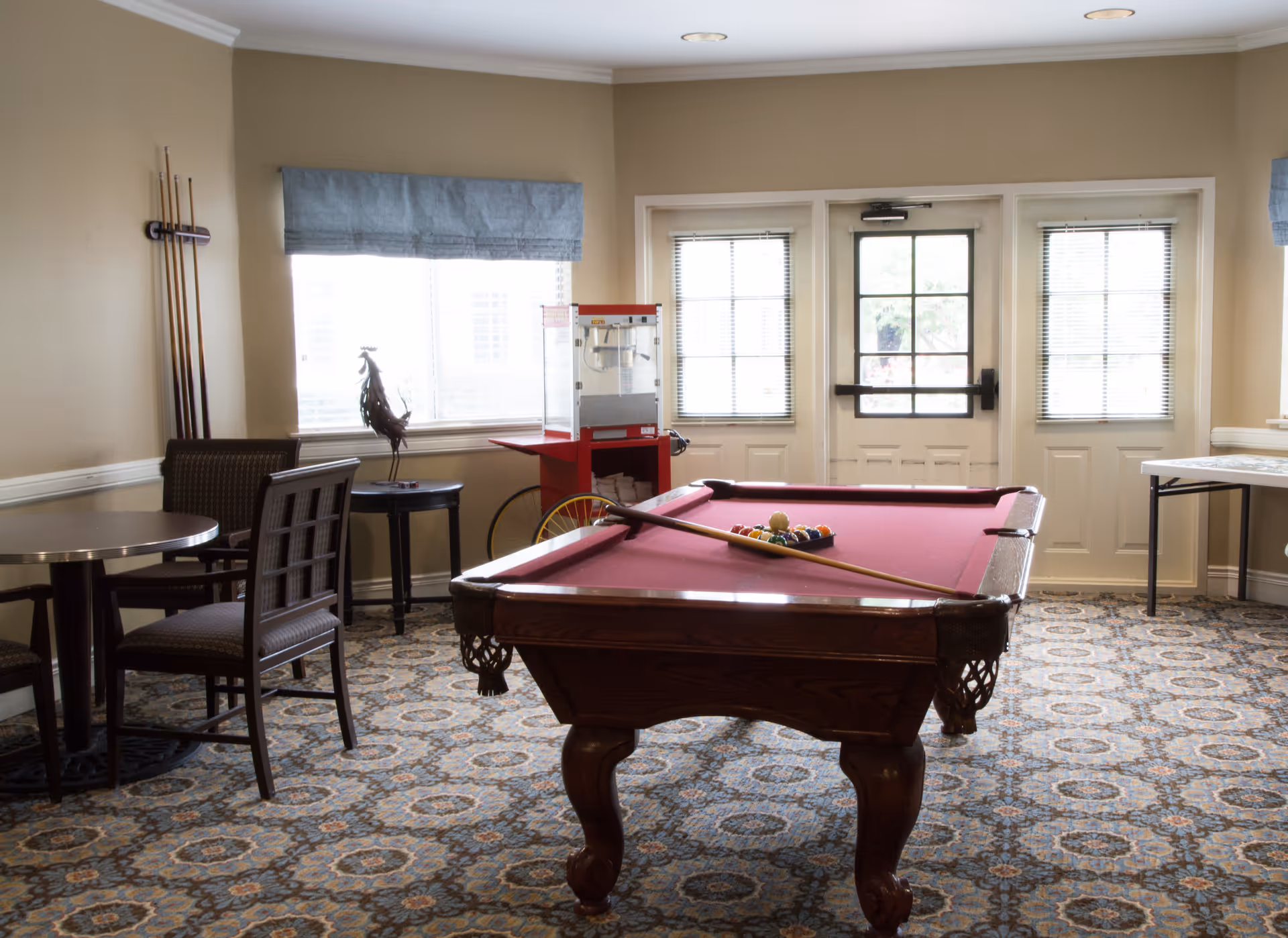 A recreational room with a red felt pool table in the center, pool cues mounted on the wall, a popcorn machine near the window, a round table with chairs, and a patterned carpet floor. The room has beige walls and windows with blue valances letting in natural light.