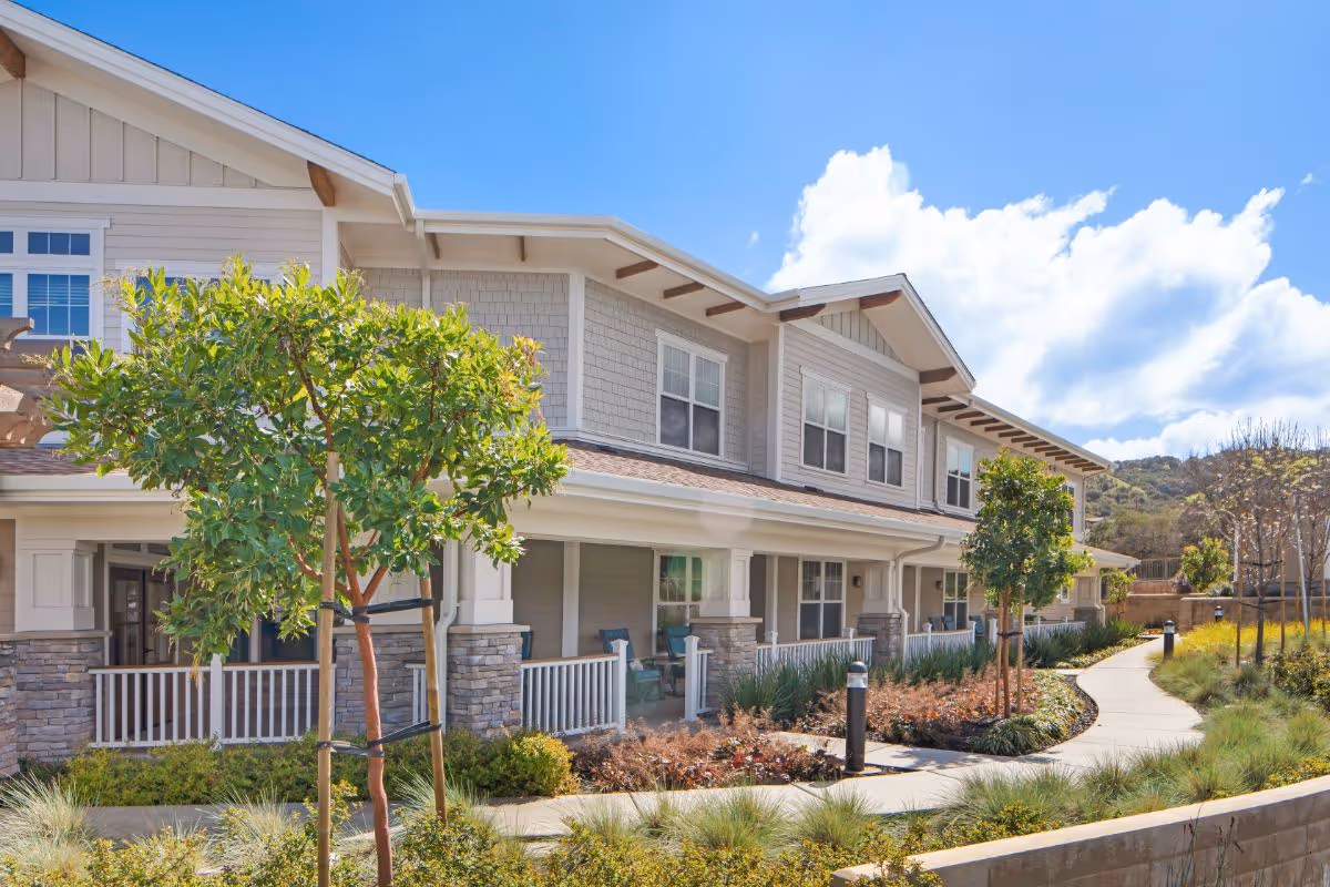 Exterior view of a two-story senior living facility building with beige siding and stone accents. The building has a covered porch with white railings and several windows. There is a curved walkway surrounded by landscaped greenery and small trees under a bright blue sky with some clouds.
