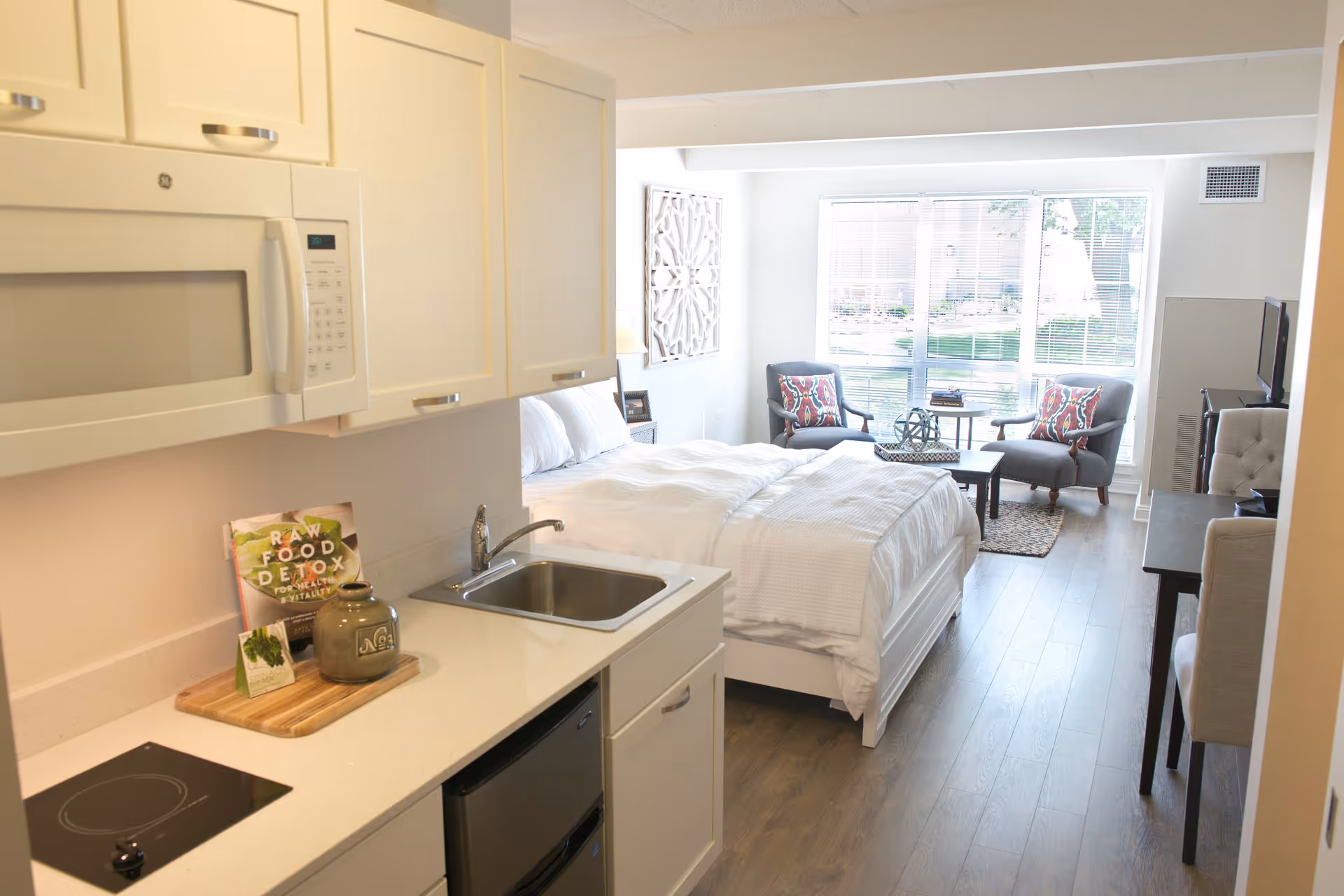 A bright studio apartment featuring a small kitchen area with white cabinets, a microwave, sink, and stovetop. The room includes a neatly made bed with white bedding, two armchairs with colorful pillows, a coffee table, and a large window letting in natural light.