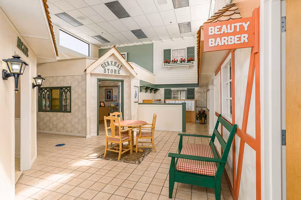 Indoor assisted-living common area designed like a small village with a 'General Store' facade, a 'Beauty Barber' sign, a table with chairs, and a green bench.