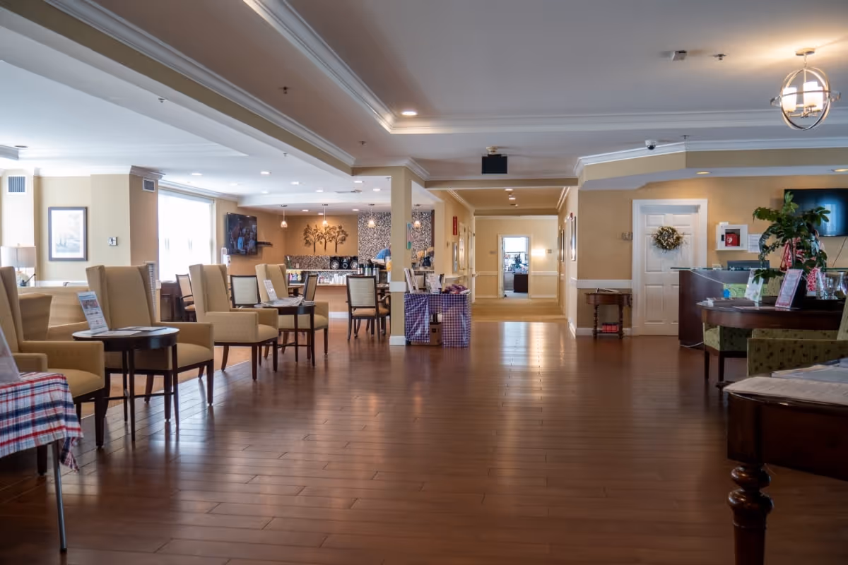 Spacious senior-living common area with rows of chairs and tables, a reception desk to the right, and a dining/service area in the background.