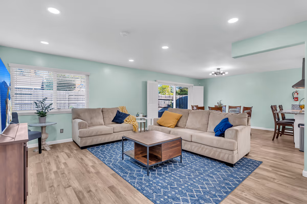 Open-plan living room with two beige sofas, a blue patterned rug, a wooden coffee table, and a dining area visible in the background.