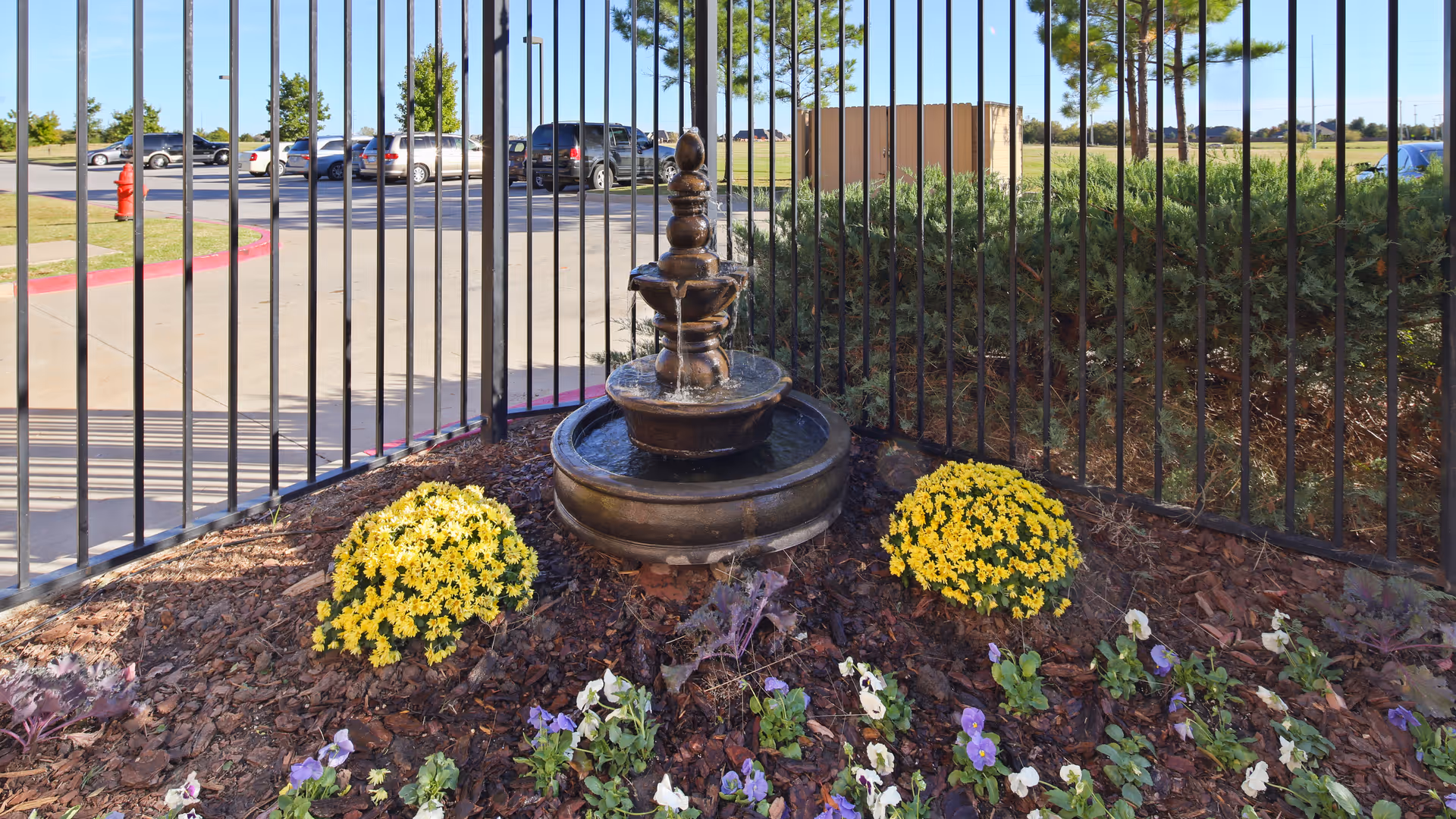 A small decorative water fountain surrounded by yellow and purple flowers, enclosed by a black metal fence with a parking lot and cars visible in the background under a clear blue sky.