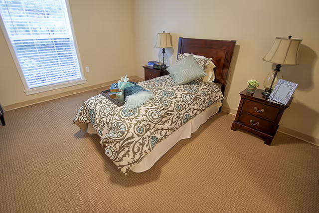 A neatly made single bed with a patterned bedspread and decorative pillows in a carpeted bedroom. There are two wooden nightstands on either side of the bed, each with a lamp. One nightstand has a small plant and a framed photo, while the other has a tray with a cup and a small decorative item. A window with blinds is on the left wall.