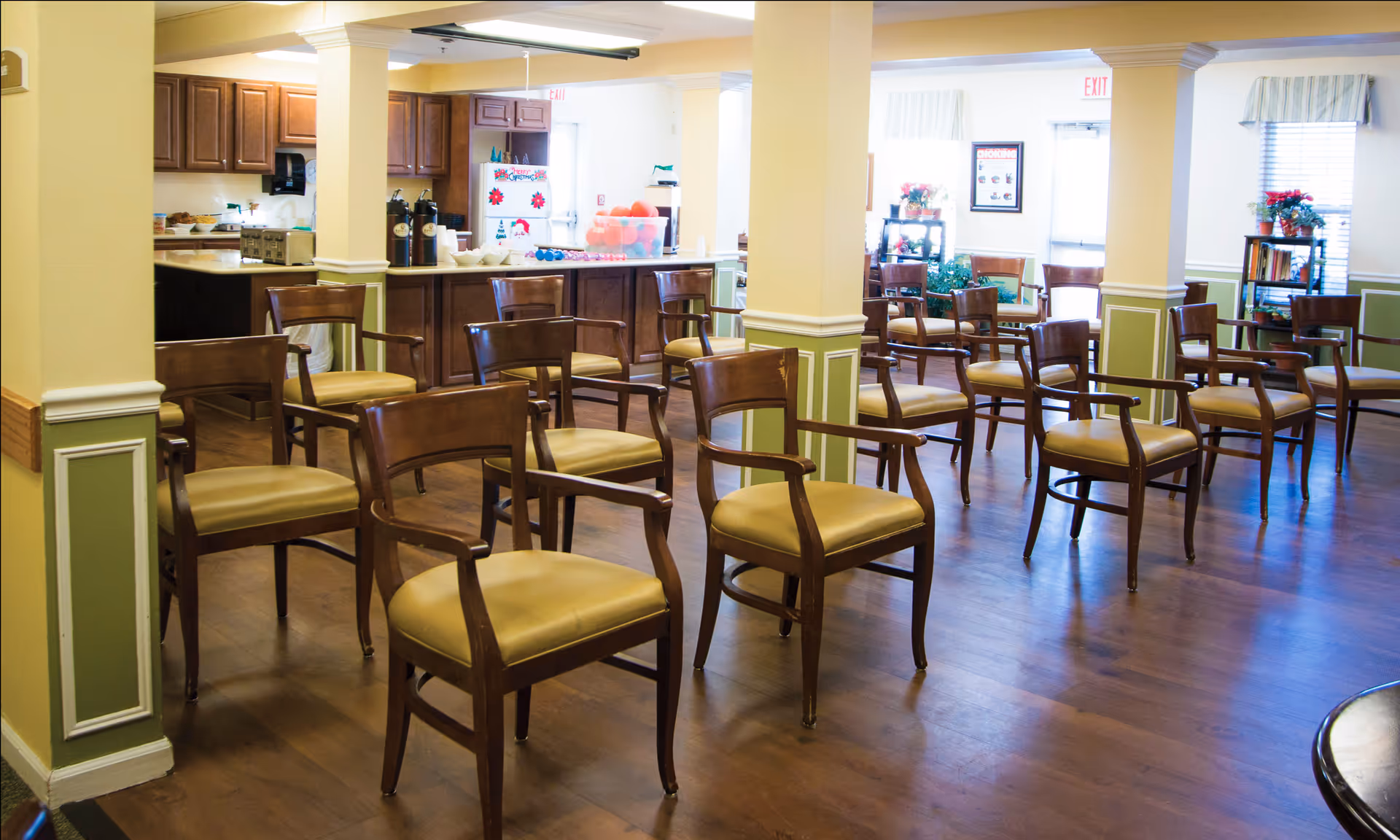 A spacious room with multiple wooden chairs arranged in rows on a wooden floor. The room has cream-colored walls with green wainscoting and white trim. In the background, there is a kitchen area with wooden cabinets, a refrigerator decorated with holiday magnets, and a counter with coffee dispensers and bowls. There are windows with striped valances and some plants and bookshelves near the walls.