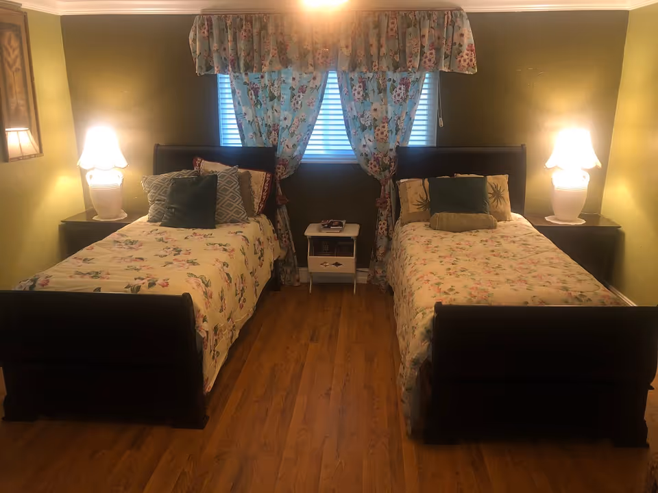 A cozy bedroom with two twin beds featuring floral bedspreads and multiple pillows. Each bed has a dark wooden frame and is flanked by a nightstand with a white lamp. Between the beds is a small white table with books on it. The room has wooden flooring, green walls, and a window with floral curtains in the center background.