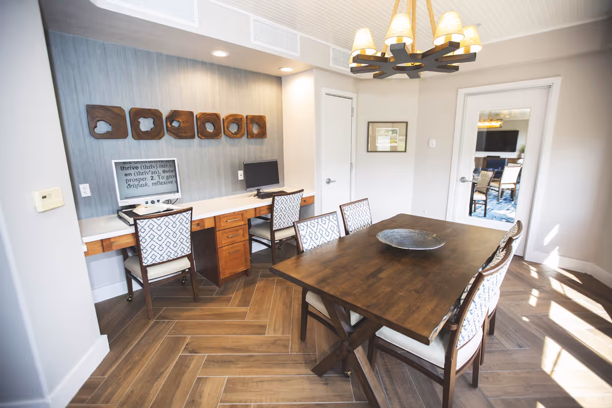 A bright room with a wooden table surrounded by six chairs with patterned upholstery. On the left side, there is a built-in desk with two chairs and two computer monitors. The wall behind the desk has decorative wooden art pieces. A modern chandelier with lampshades hangs above the table. There is a door with a glass panel leading to another room.