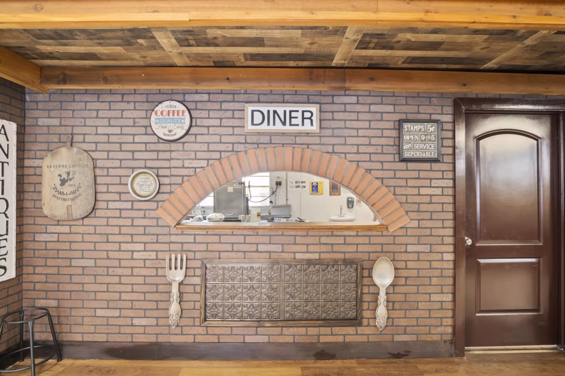 Interior view of a brick wall with a small arched window opening into a kitchen area. The wall is decorated with vintage-style signs including one that says 'DINER', a large fork and spoon, and other decorative plates and signs. There is a dark wooden door to the right and a wooden stool to the left.
