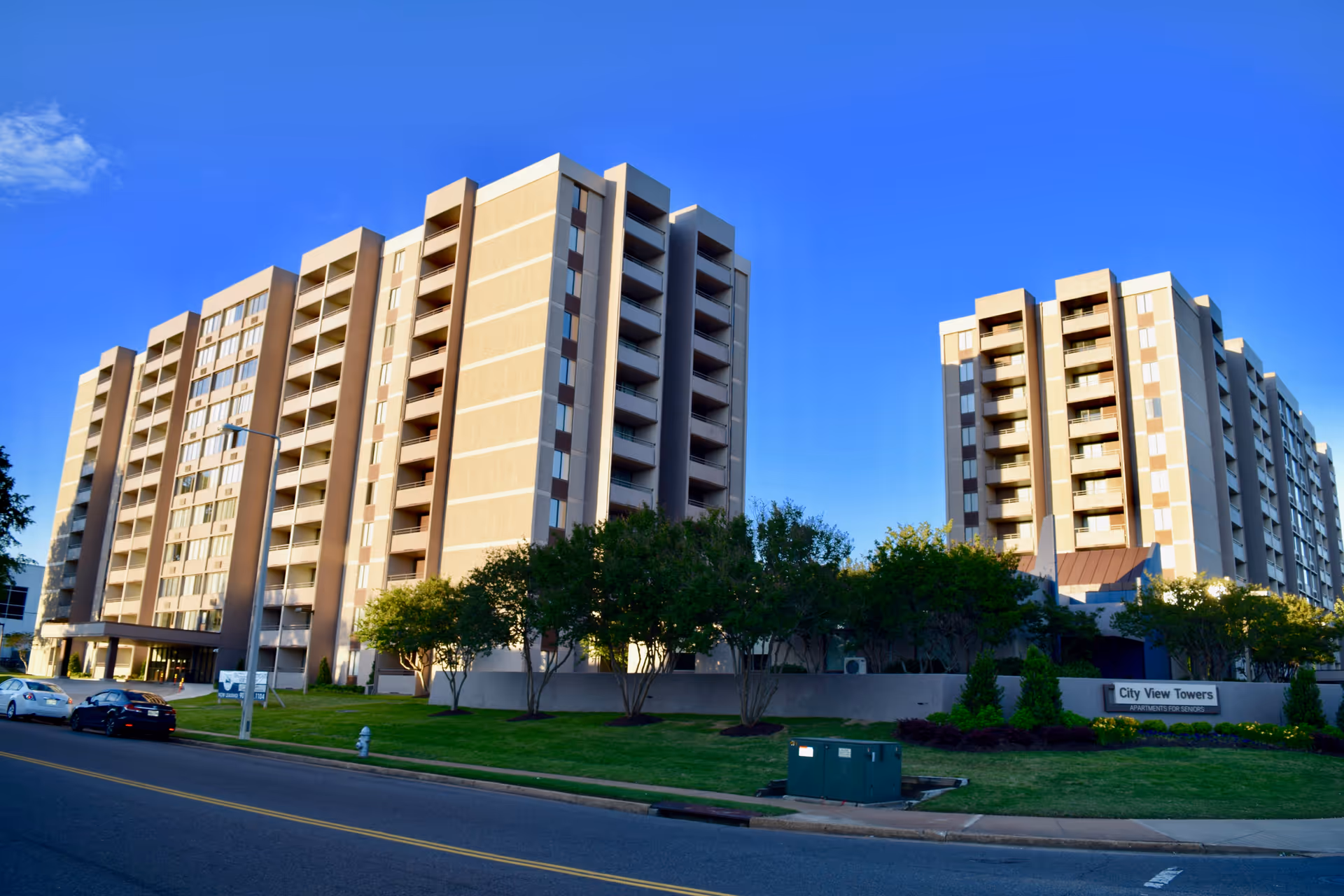 Exterior view of City View Towers Senior Apartments, showing two multi-story residential buildings with balconies, surrounded by trees and a well-maintained lawn under a clear blue sky.