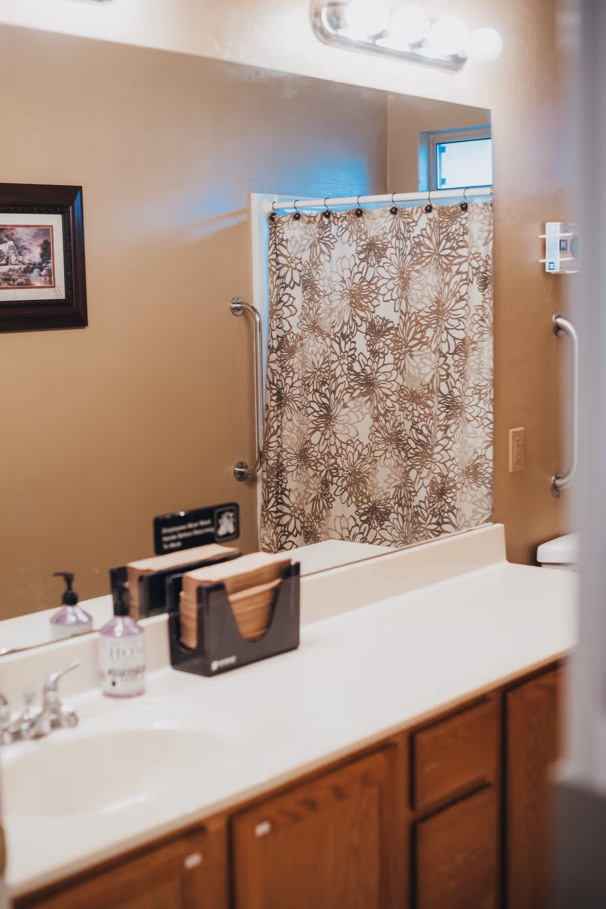 Bathroom vanity with a sink and mirror, floral shower curtain, grab bars, and toiletries on the countertop.