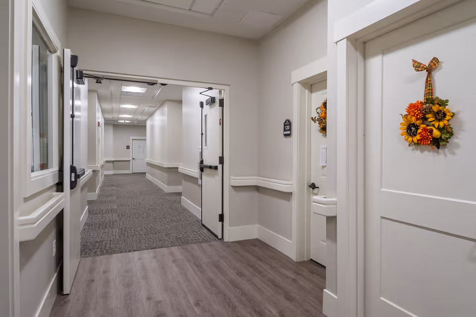 Interior hallway of a senior living facility with light-colored walls and flooring. The hallway has open double doors leading to a carpeted corridor with additional closed doors. One door on the right side is decorated with a fall-themed wreath featuring sunflowers and orange flowers.