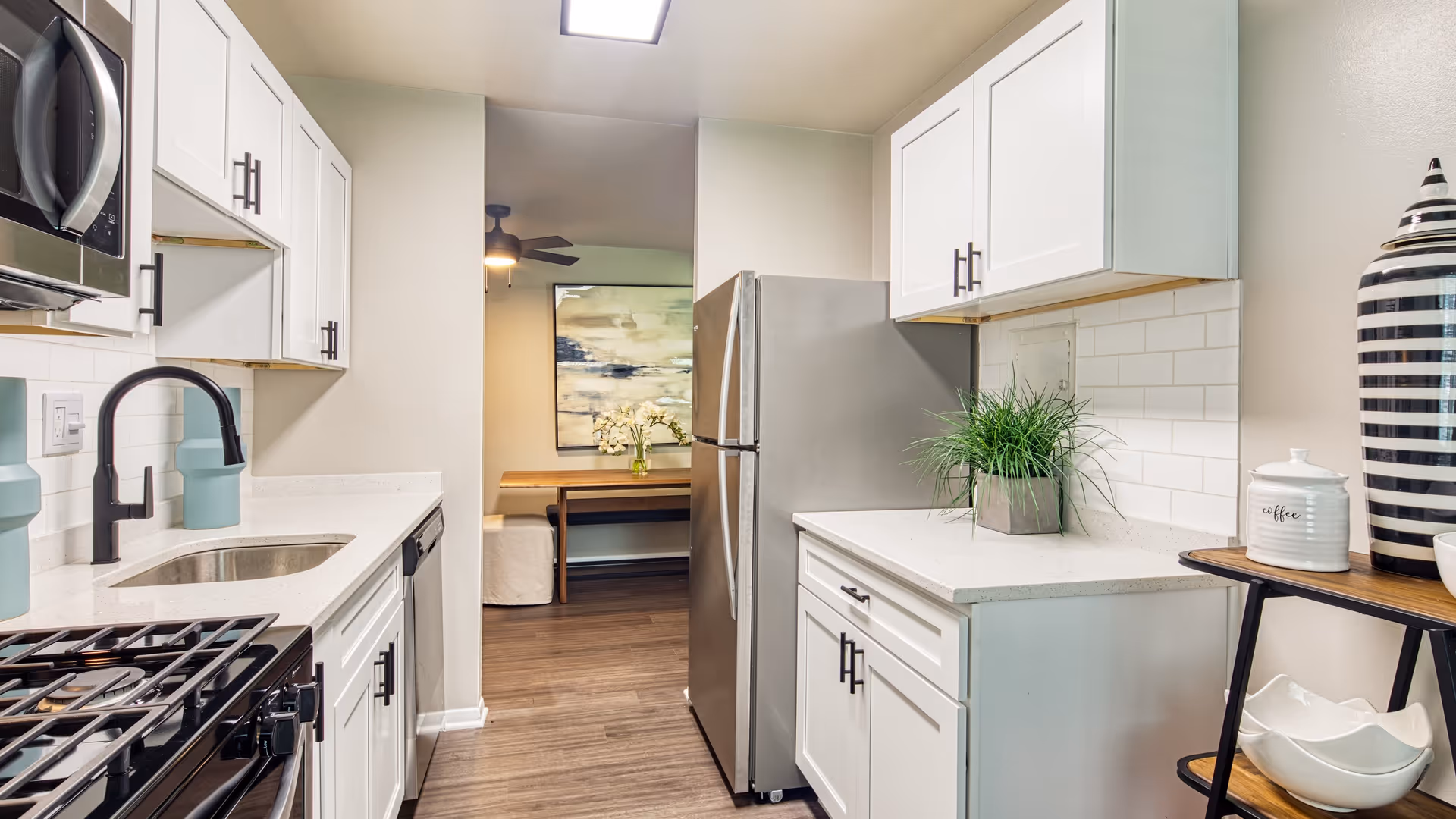 Modern kitchen with white cabinets, stainless steel appliances including a refrigerator, microwave, and stove. The kitchen has a black faucet, white countertops, and a small green plant on the counter. In the background, there is a dining area with a wooden table, bench seating, a ceiling fan, and a large abstract painting on the wall.