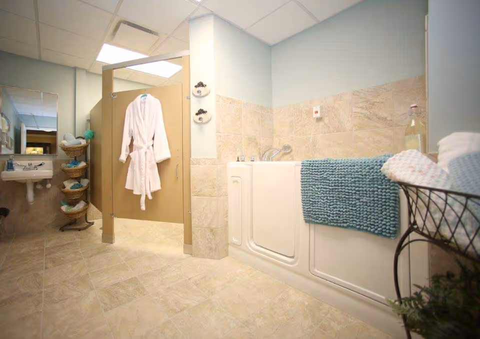 A clean and well-lit bathroom featuring a walk-in bathtub with a blue bath mat draped over the side. A white bathrobe hangs on the door of a beige shower stall. There is a small sink with a mirror above it and a tiered basket stand holding towels and bath items. The walls are tiled halfway up with beige tiles and painted light blue above.