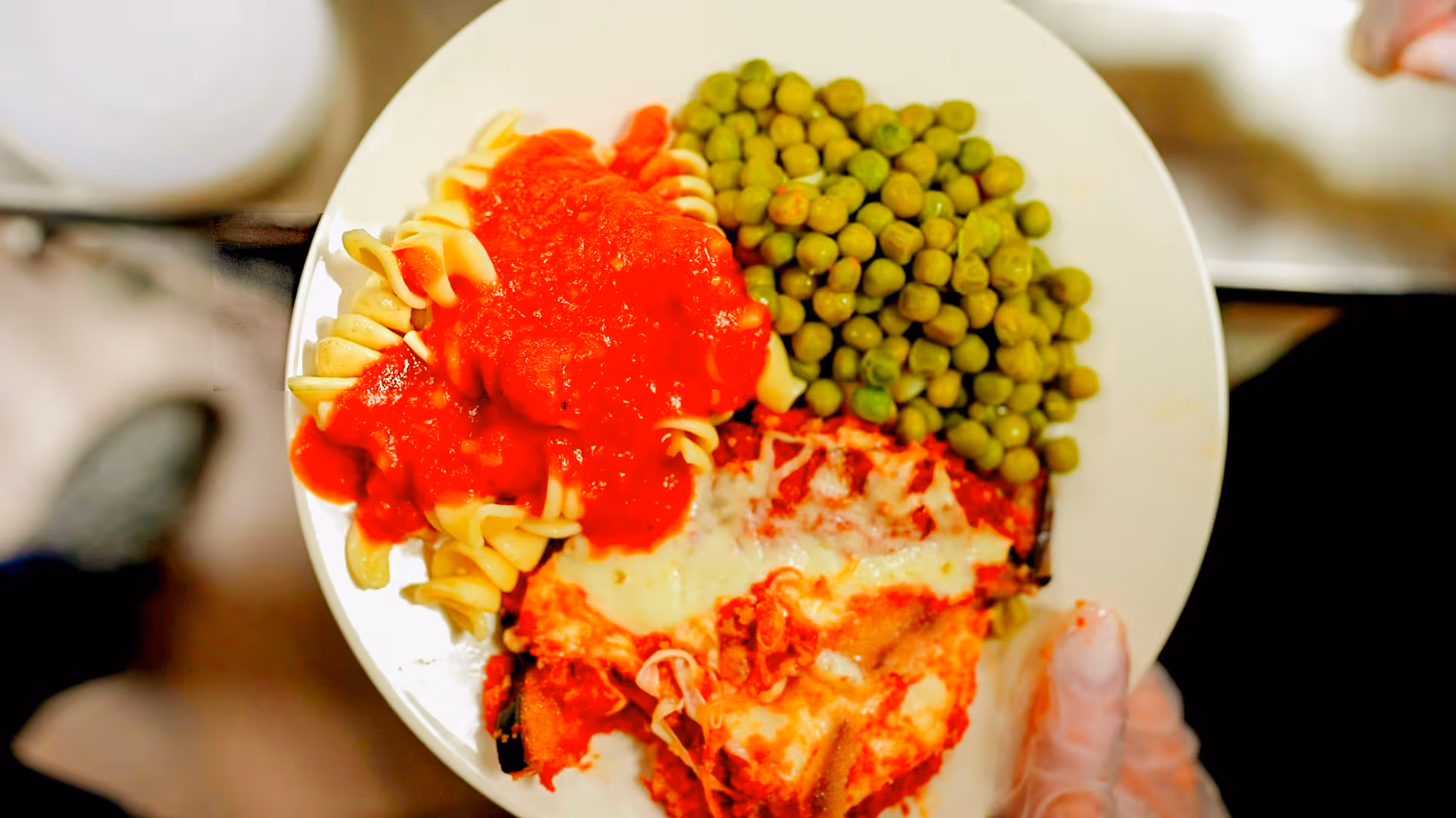 A white plate holding rotini pasta with tomato sauce, green peas, and a cheesy baked entrée being held by a gloved hand.