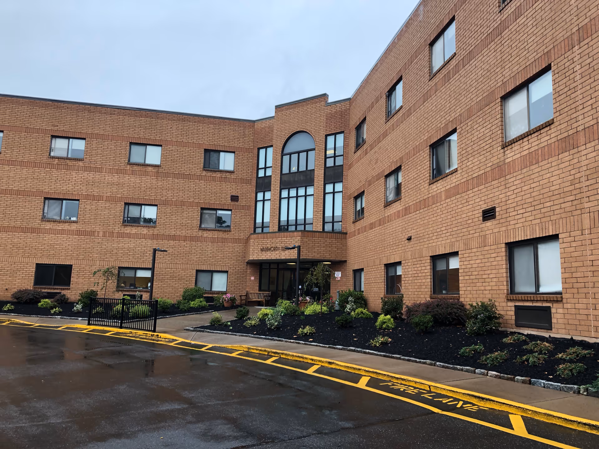 Exterior view of a three-story brick building with multiple windows and a main entrance in the center. There are landscaped bushes and plants along the front, a wet driveway with yellow painted curb markings, and a cloudy sky overhead.