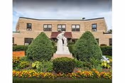 Brick building exterior with statue, manicured shrubs, and colorful flower beds in front.