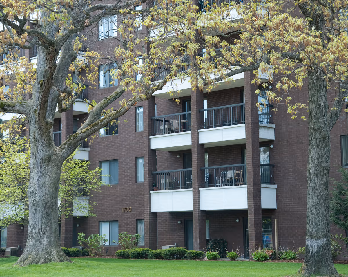 Exterior view of a multi-story brick building with balconies, surrounded by green grass and large trees with budding leaves.