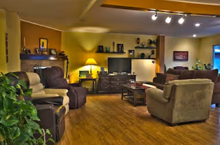 Cozy living room with multiple recliners and sofas arranged around a TV and coffee table, warm yellow lighting and plants.