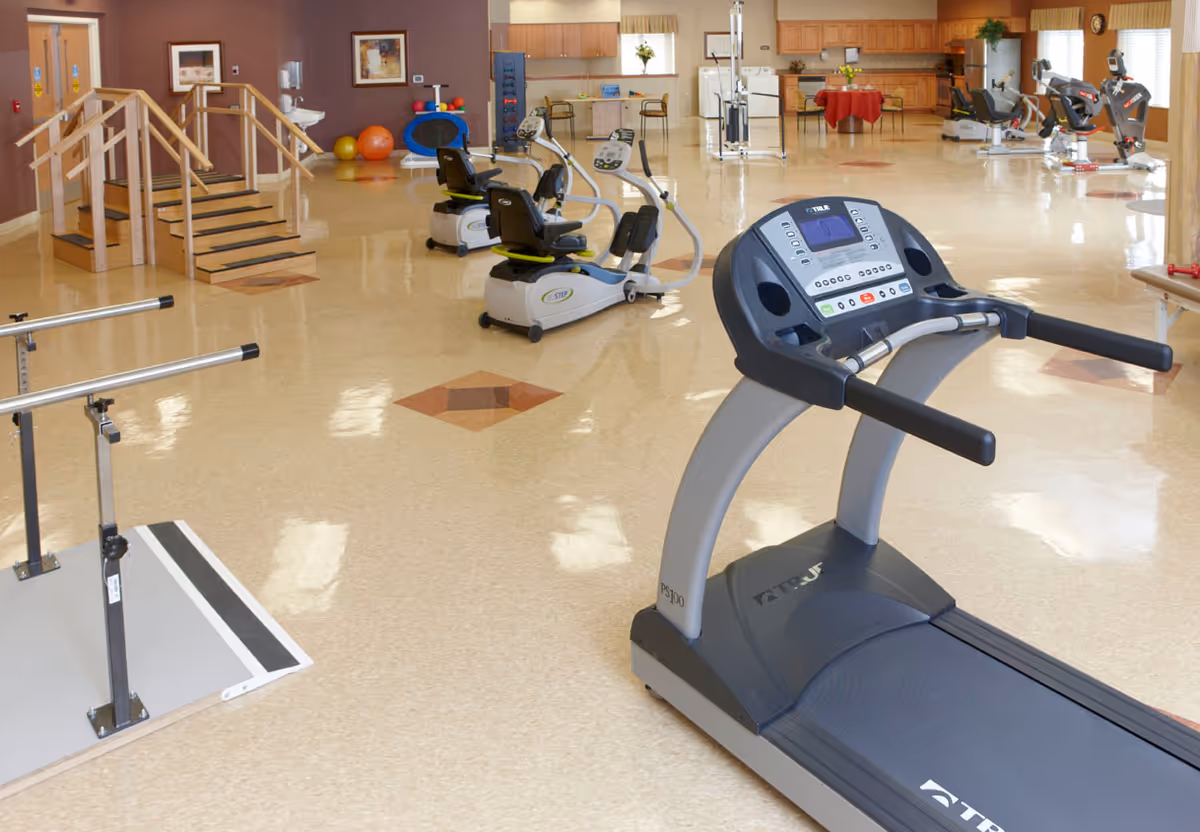 A spacious fitness and rehabilitation room with exercise equipment including a treadmill, recumbent bikes, parallel bars, and a small staircase for physical therapy. The room has a polished floor, warm lighting, and a kitchenette area with a table and chairs in the background.
