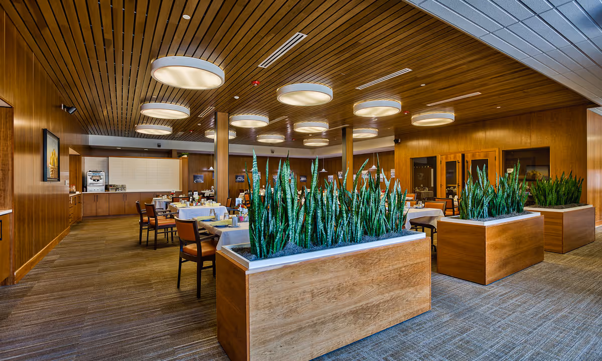A spacious dining room with wooden walls and ceiling, featuring round ceiling lights. Several tables are set with white tablecloths and chairs arranged around them. Large wooden planters with tall green plants divide the space. In the background, there is a counter area with a coffee machine and cabinets.