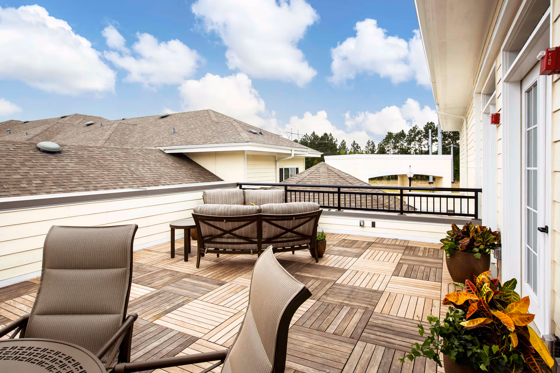 Outdoor patio area with wooden patterned flooring, cushioned seating, a small table, potted plants, and a view of rooftops and trees under a partly cloudy sky.