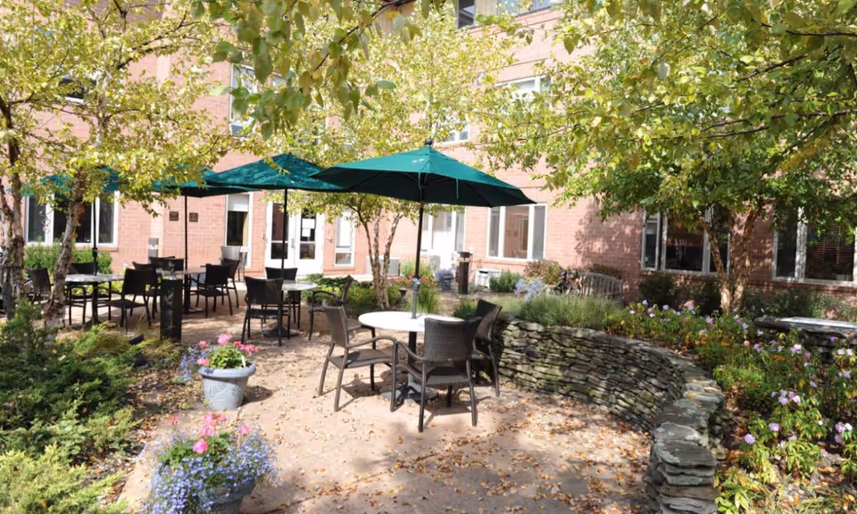 Outdoor patio area at Daughters of Miriam Center for Nursing and Rehabilitation with round tables, chairs, green umbrellas, surrounded by trees, plants, and flowers, adjacent to a brick building.
