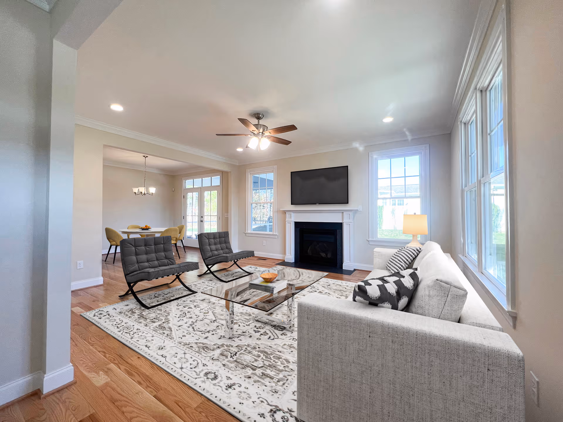Bright modern living room with a fireplace and wall-mounted TV, a gray sofa, two black chairs, coffee table, and large windows.