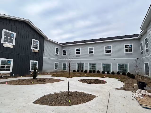 Outdoor courtyard area of a senior living facility under construction with newly planted small trees and shrubs surrounded by concrete walkways, and a two-story building with multiple windows in the background.