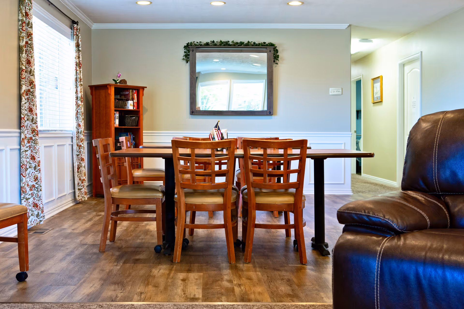 A dining area with a wooden table surrounded by six wooden chairs with cushioned seats. There is a large mirror on the wall above the table, a wooden bookshelf with books and baskets in the corner, and a window with floral curtains letting in natural light. Part of a brown leather armchair is visible on the right side, and a hallway with doors and a framed picture is seen in the background.
