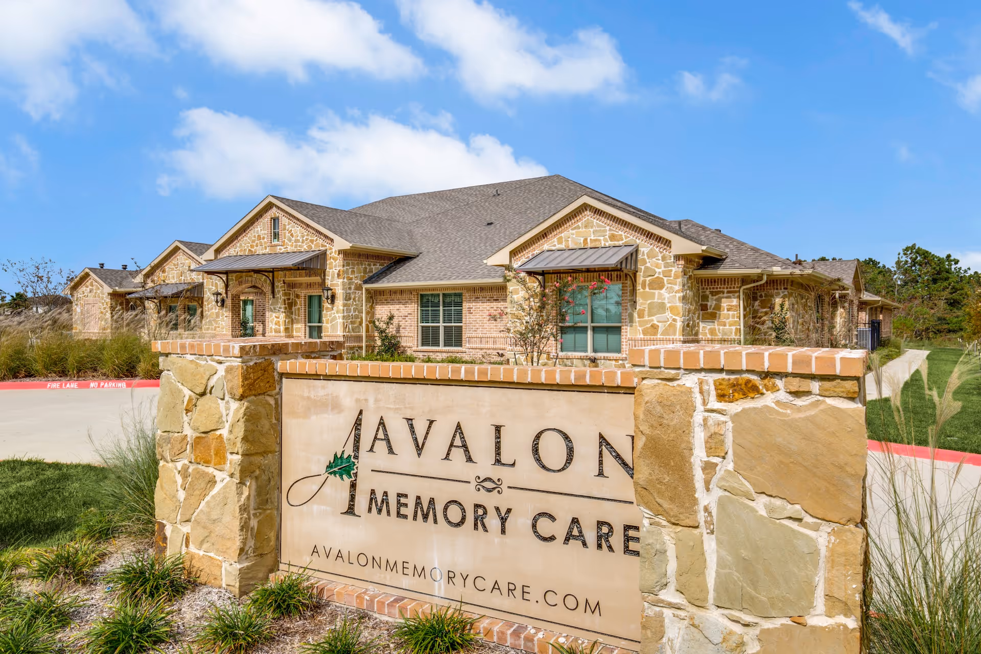 Exterior view of Avalon Memory Care facility showing a stone and brick building under a blue sky with some clouds. In the foreground, there is a large stone sign with the facility name and website.
