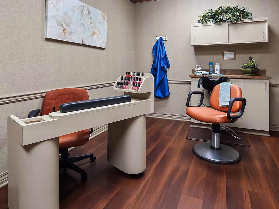 A small salon room with a manicure table and chair on the left, a display of nail polish bottles, and a hair washing station with an orange salon chair and a towel draped over it. The room has wood flooring, beige walls, a floral painting, and a blue salon cape hanging on the wall.