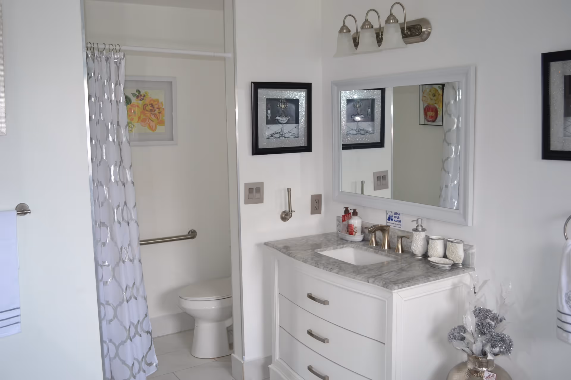 A clean and bright bathroom featuring a white vanity with a marble countertop, a large mirror with three light fixtures above it, a toilet partially visible behind a shower curtain with a geometric pattern, and decorative framed artwork on the walls.