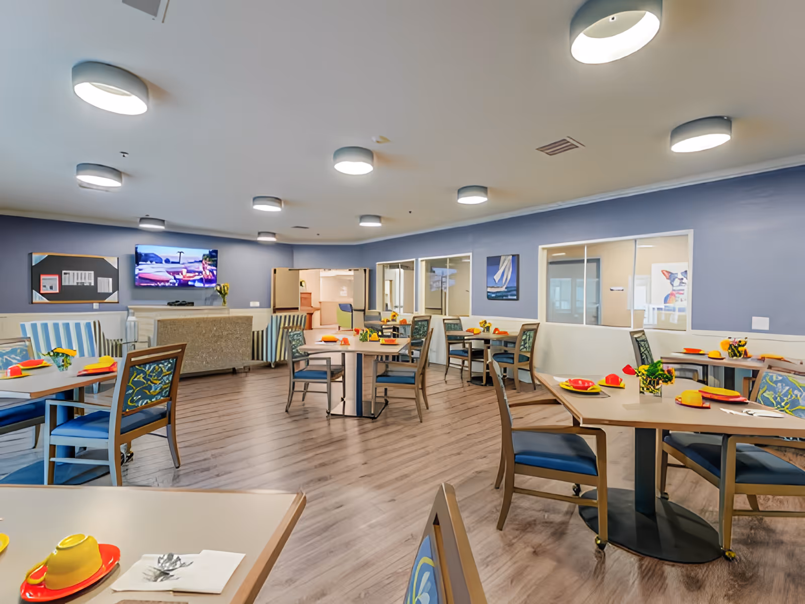 A bright and spacious dining room with several tables and chairs arranged neatly. Each table is set with colorful plates, bowls, and napkins, and small flower arrangements. The room has light wooden flooring, blue walls, and multiple ceiling lights. A flat-screen TV is mounted on one wall above a fireplace, and there are paintings and windows on the walls.