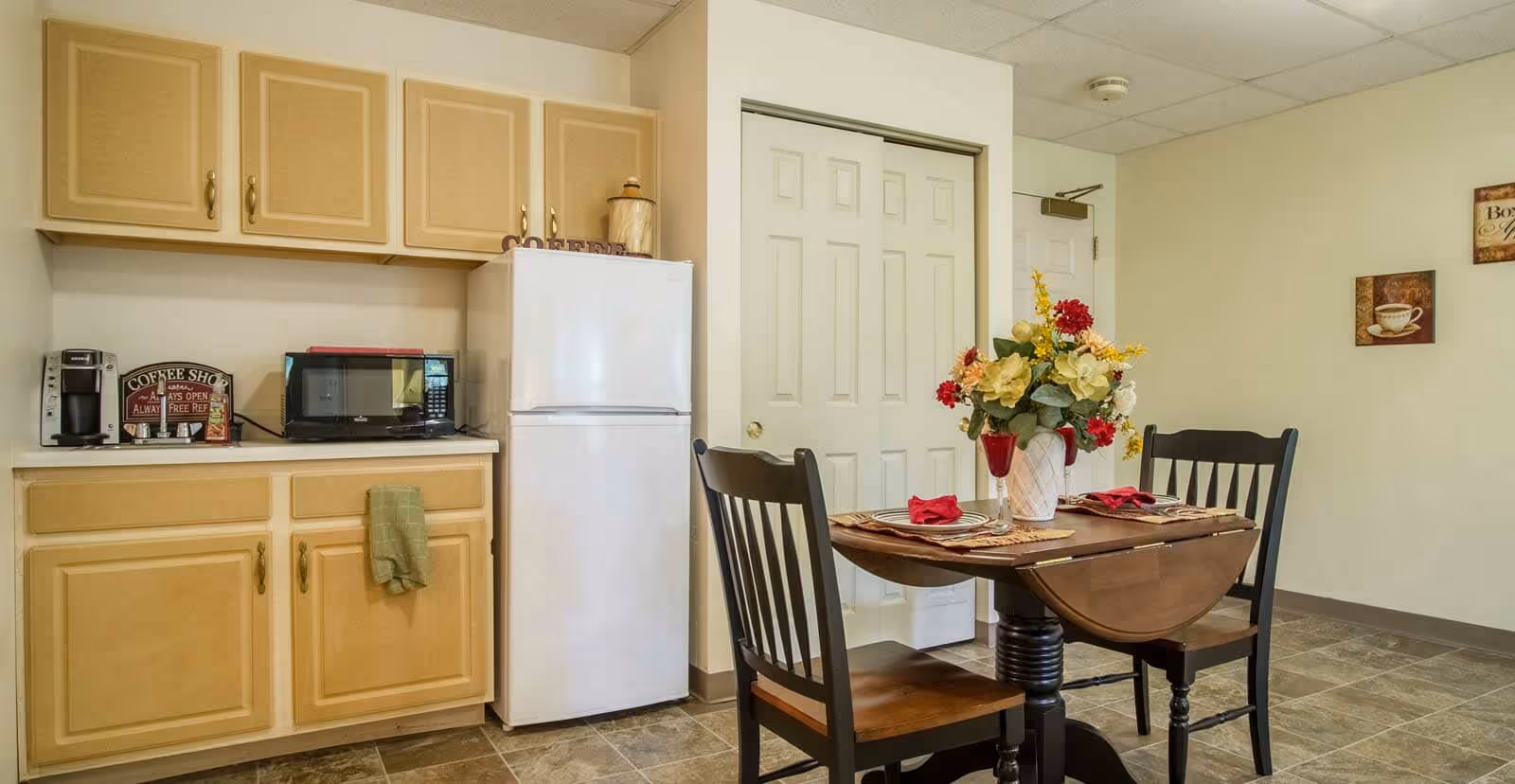 A small kitchen area with beige cabinets, a white refrigerator, a microwave, and a coffee maker on the countertop. Next to the kitchen is a wooden dining table set for two with a floral centerpiece and red napkins. The room has tiled flooring and light-colored walls with coffee-themed wall art.