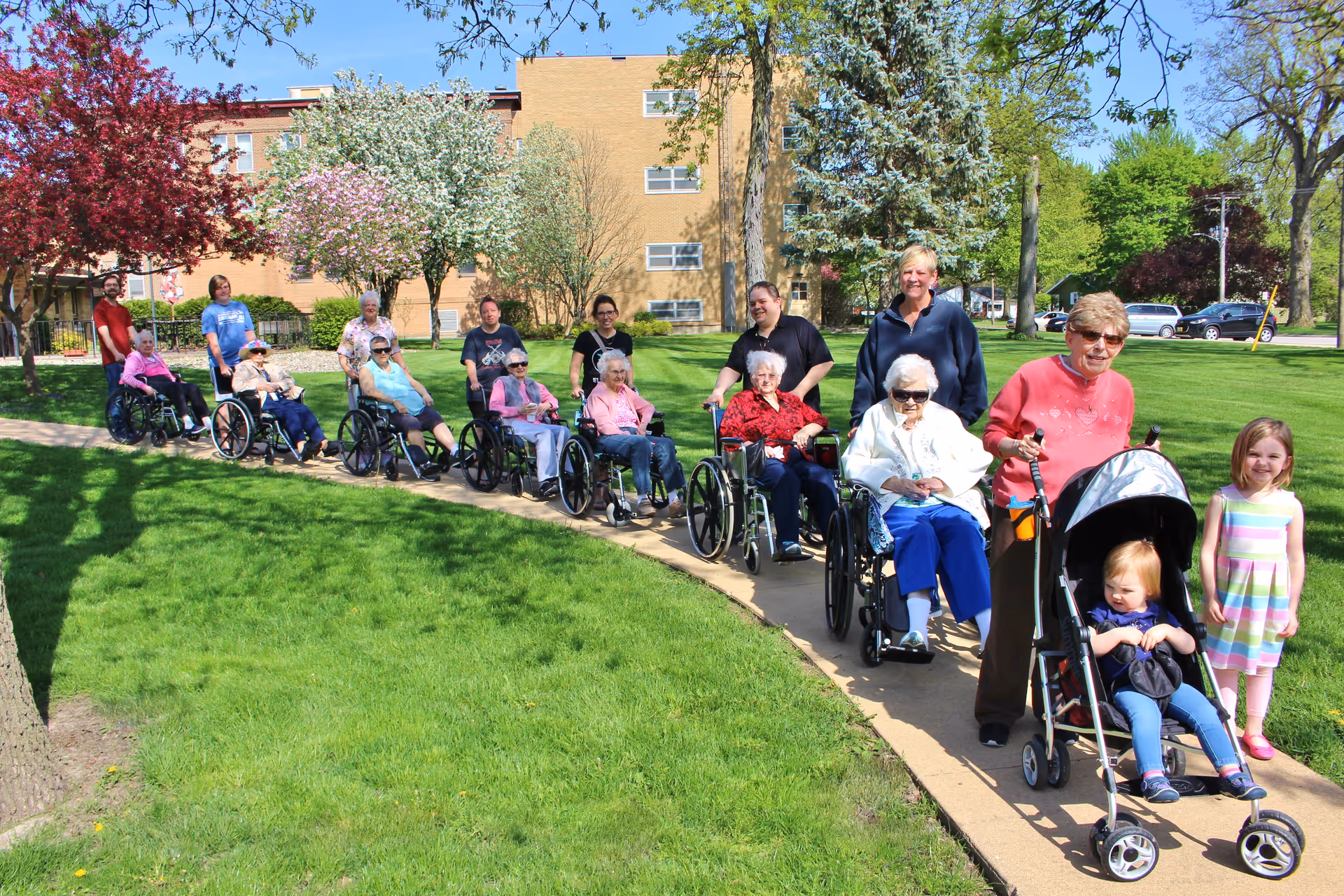 A group of elderly people, many in wheelchairs, are outside on a sunny day on a paved path in a green grassy area with trees and a building in the background. Several caregivers and two young children, one in a stroller, accompany them. The scene shows a cheerful outdoor outing at a senior living facility.