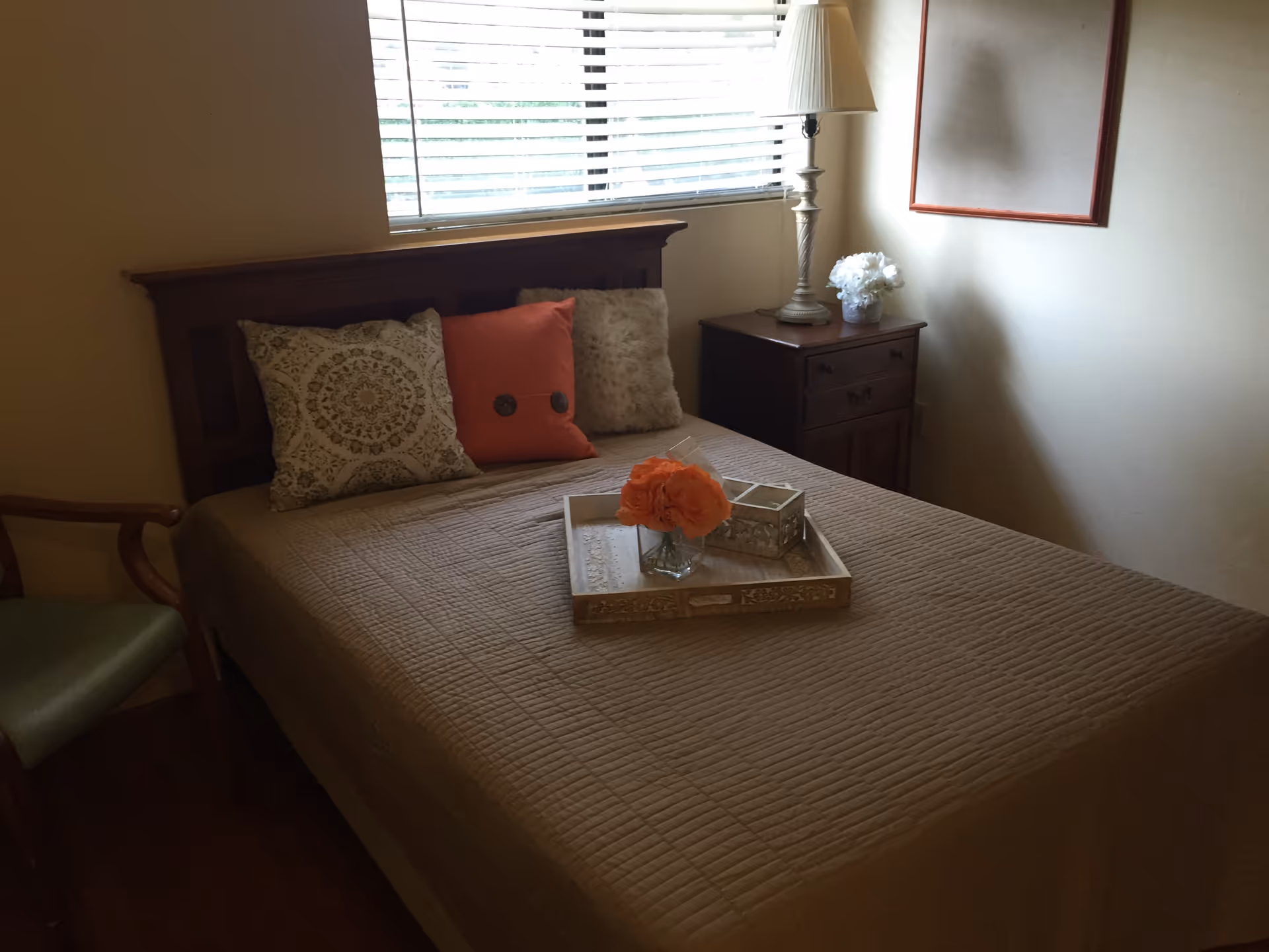 A bedroom with a bed covered in a beige quilt and three decorative pillows in beige, orange, and furry textures. A wooden nightstand with a lamp and a small flower arrangement sits next to the bed. A wooden chair with a green cushion is partially visible on the left side. A window with white blinds is above the bed, and a framed empty board hangs on the wall.