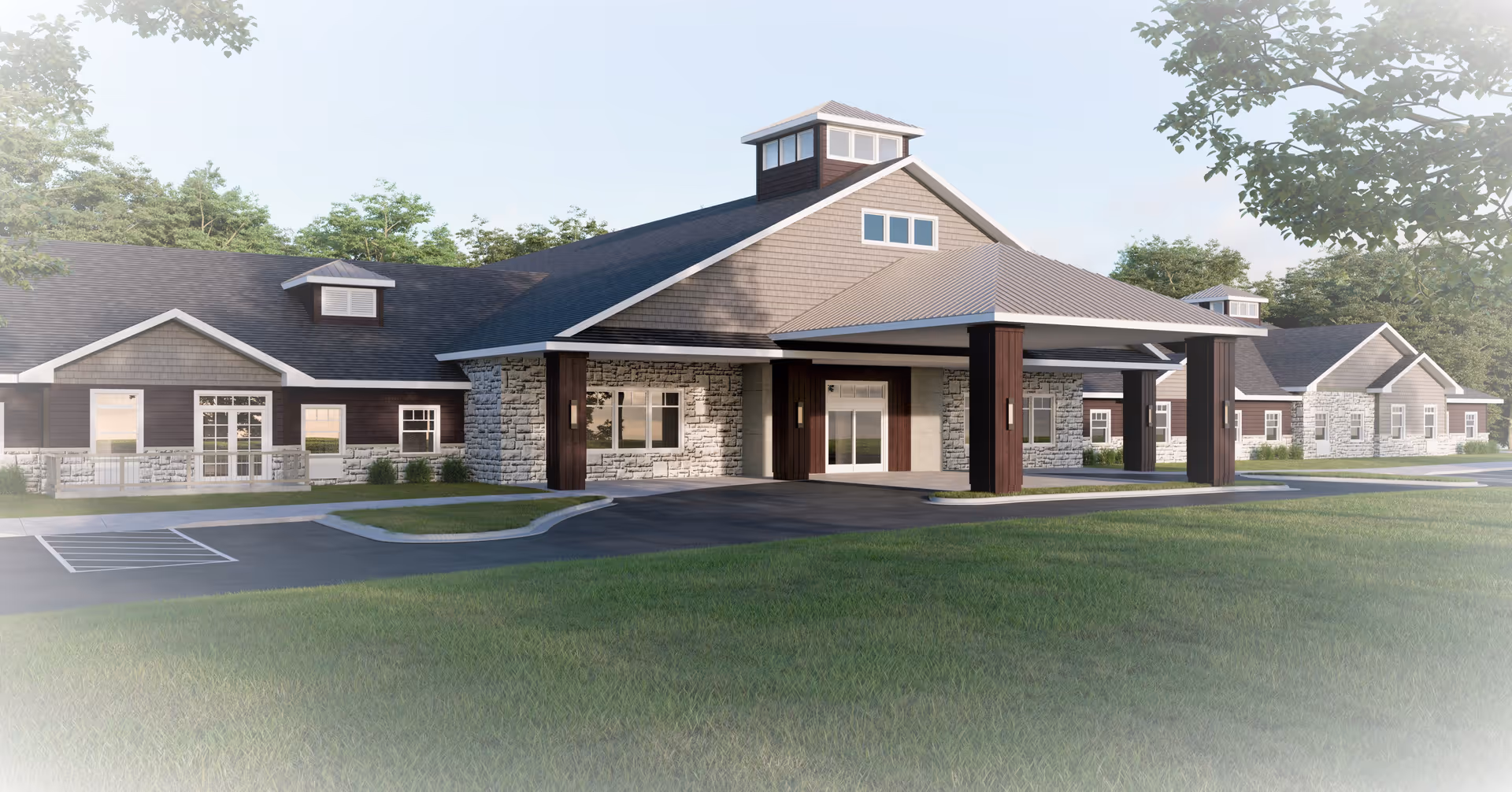 Exterior view of a single-story senior living facility building with a covered entrance supported by four dark wooden pillars. The building features a combination of stone and dark siding with multiple windows and a pitched roof with small dormer windows. The surrounding area includes a paved driveway, parking spaces, and green lawn with trees in the background under a clear sky.