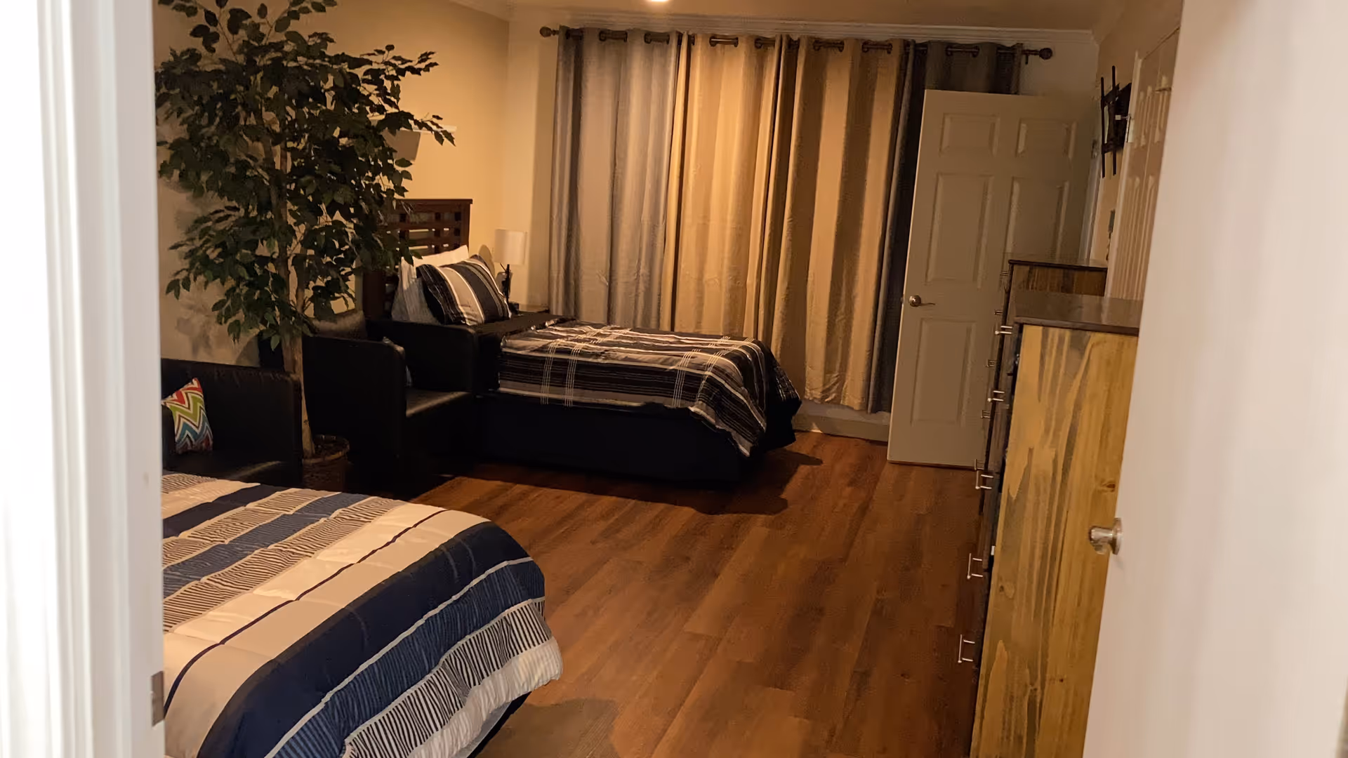 Spacious bedroom with two beds, wood floors, a dresser, armchair, potted plant, and drawn curtains.