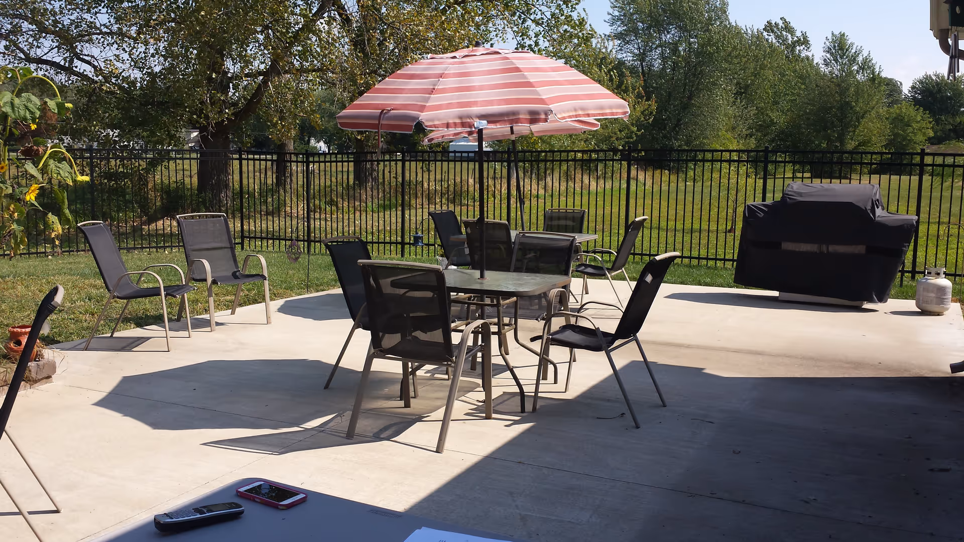 Outdoor patio with a round glass table, several chairs, a red-striped umbrella and a covered grill beside a fenced grassy area.