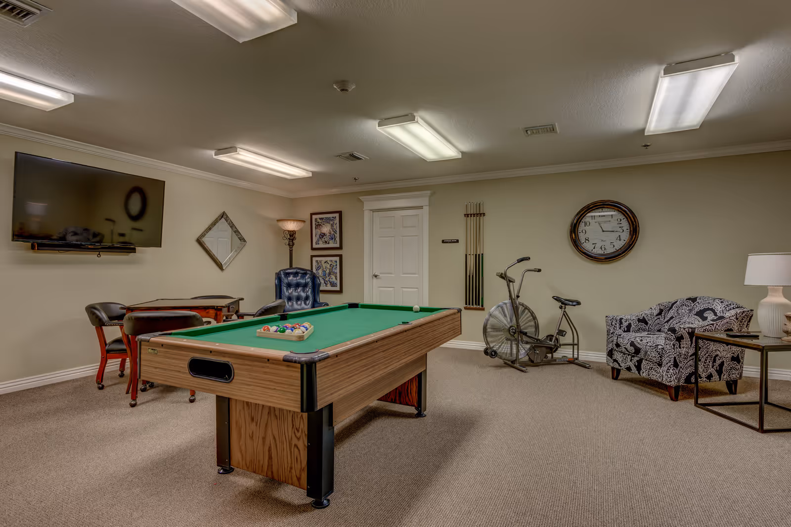 A recreational room in a senior living community featuring a pool table with balls arranged for a game, a stationary exercise bike, a patterned armchair next to a side table with a lamp, a wall clock, a flat-screen TV mounted on the wall, a small table with four chairs, and framed artwork on the walls.