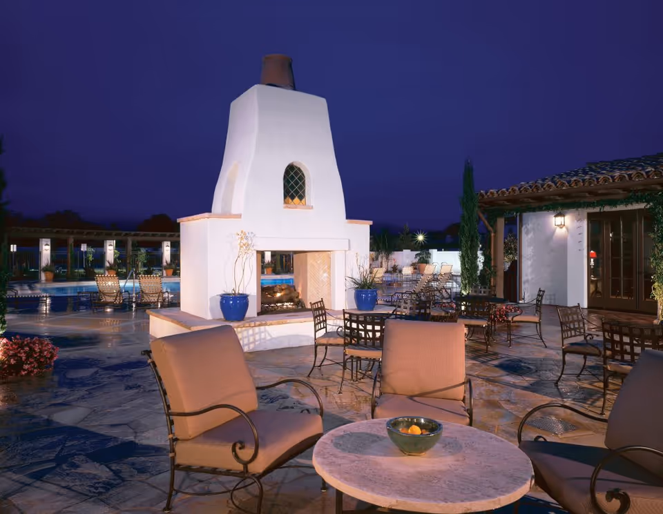 Outdoor patio area at night with cushioned chairs and round tables surrounding a large white outdoor fireplace. There are potted plants, lounge chairs near a pool, and a building with a tiled roof and lit wall lantern in the background.