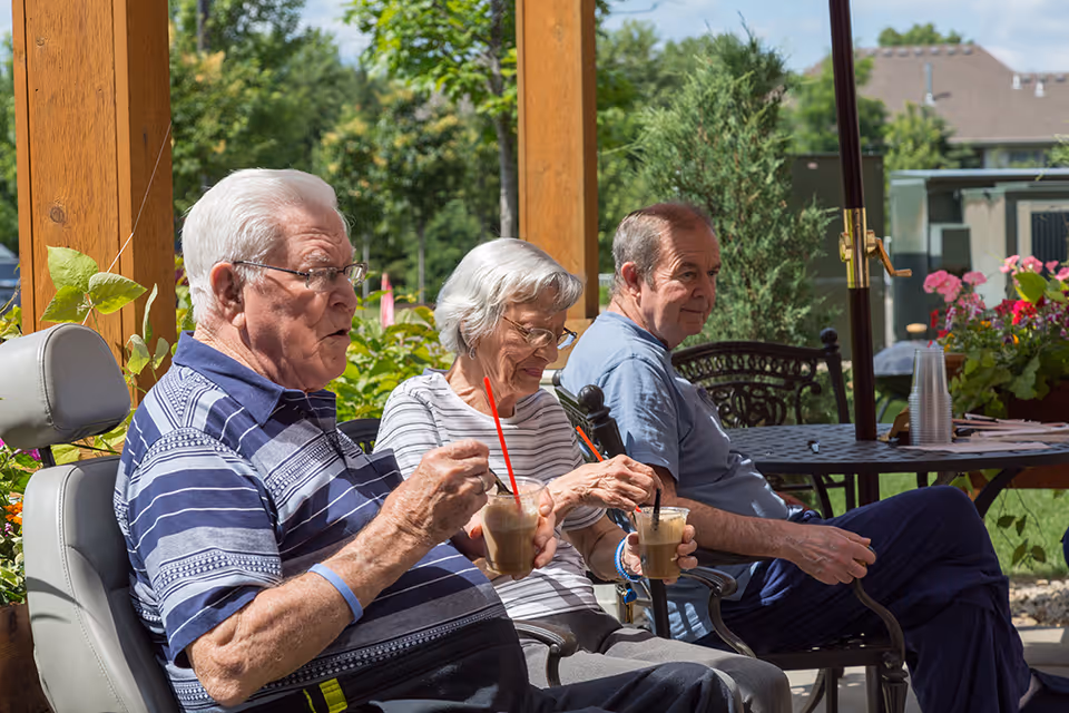 Three elderly individuals sitting outdoors on a patio, enjoying iced beverages with straws. They are seated on chairs under a wooden pergola with greenery and flowers in the background.