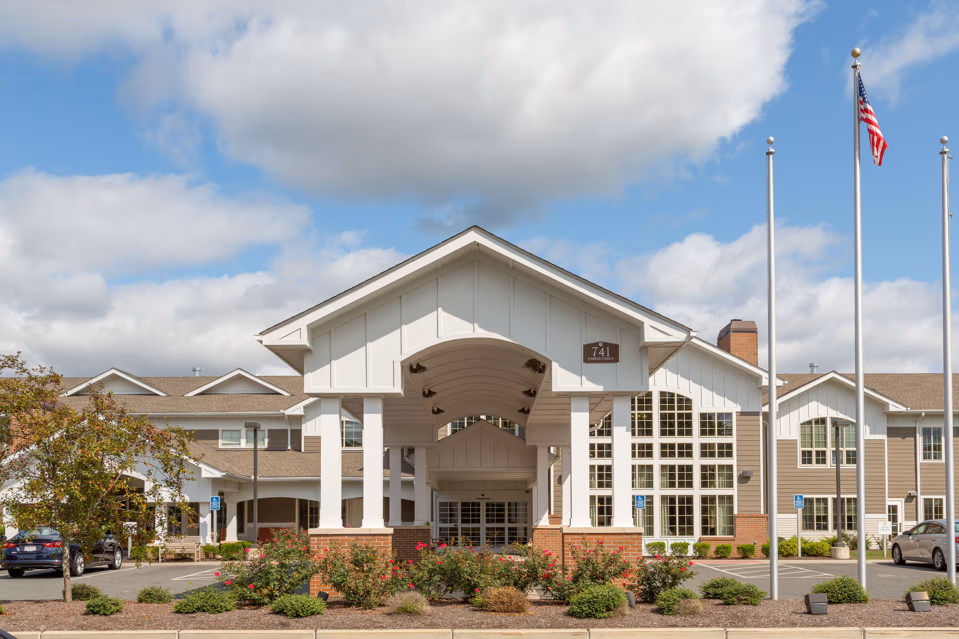 Front exterior view of The Reserve at East Longmeadow building with a covered entrance, landscaped bushes, and an American flag on a flagpole under a partly cloudy sky.