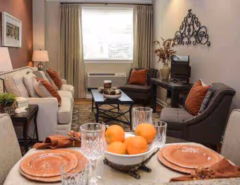Cozy living room with a white sofa, two gray armchairs with orange cushions, a black coffee table with decorative items, a TV on a black stand, and a window with beige curtains. In the foreground, a dining table is set with orange plates, crystal glasses, and a bowl of oranges.