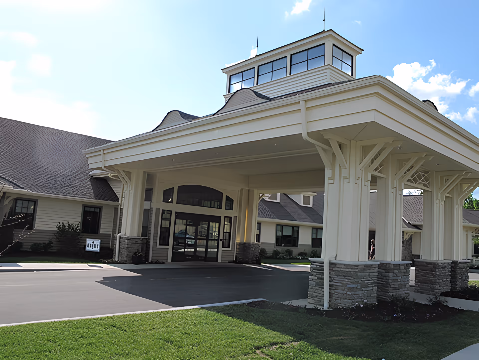 Entrance of a senior living facility with a large covered driveway supported by white columns with stone bases, beige siding, and a multi-windowed cupola on the roof under a blue sky with some clouds.