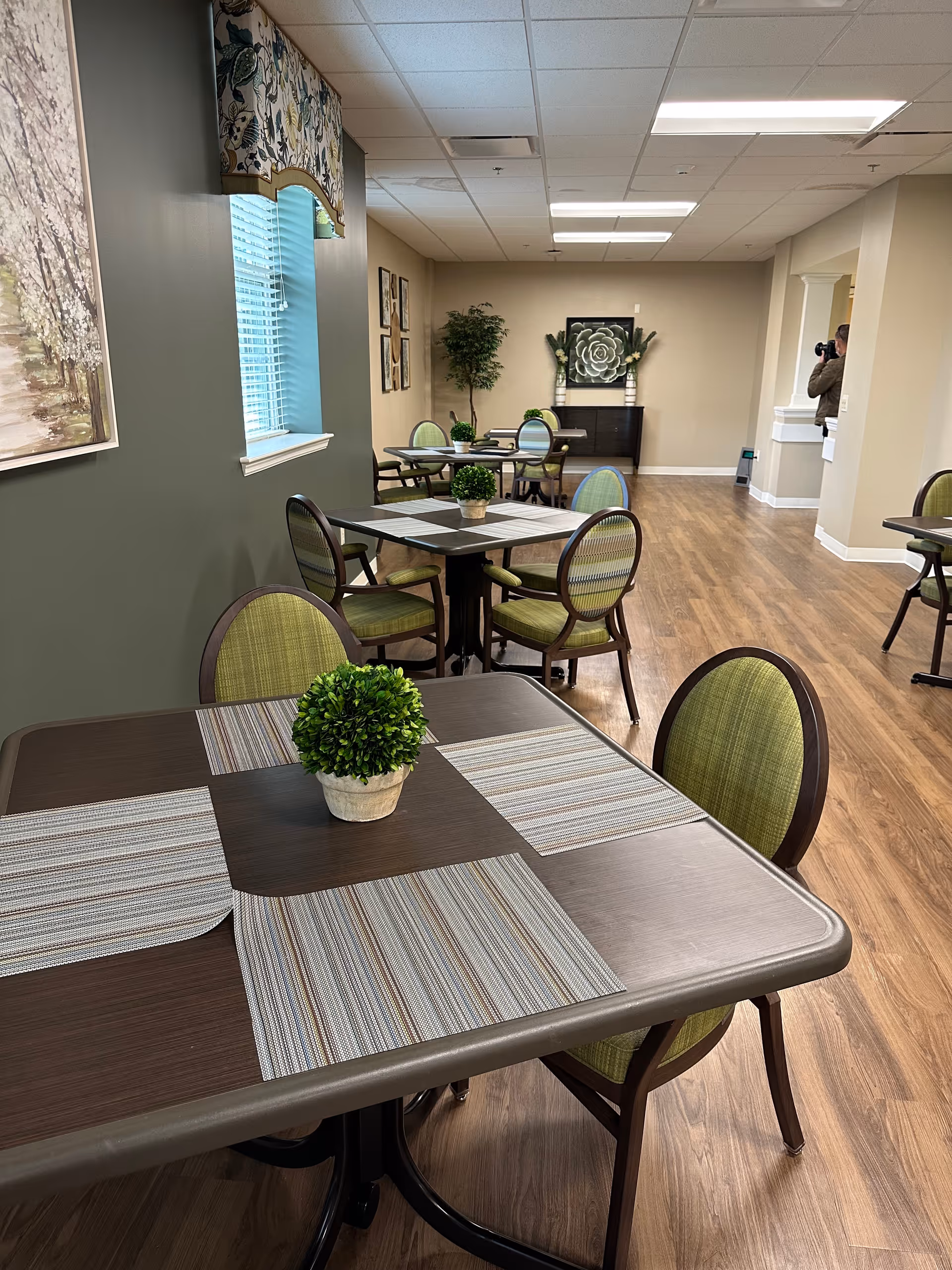 Interior view of a dining area in a senior living facility with several tables and green cushioned chairs. Each table has placemats and a small potted plant centerpiece. The room has wood flooring, beige walls, and decorative artwork on the walls. A person with a camera is partially visible in the background near a hallway.