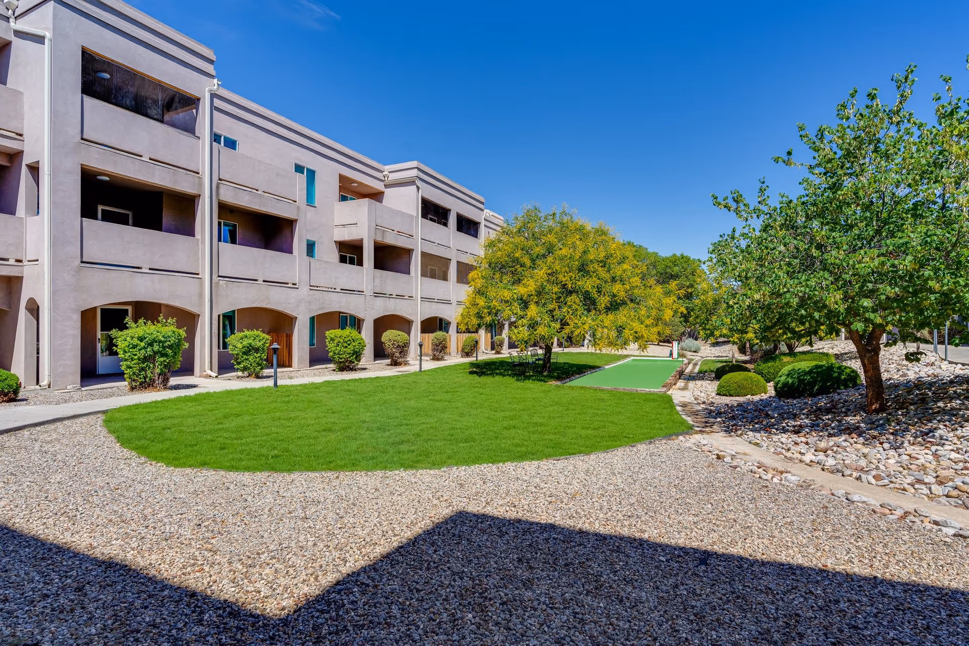 Outdoor view of Bear Canyon Estates showing a beige multi-story building with balconies, a green lawn area, trees, bushes, and a bocce ball court under a clear blue sky.