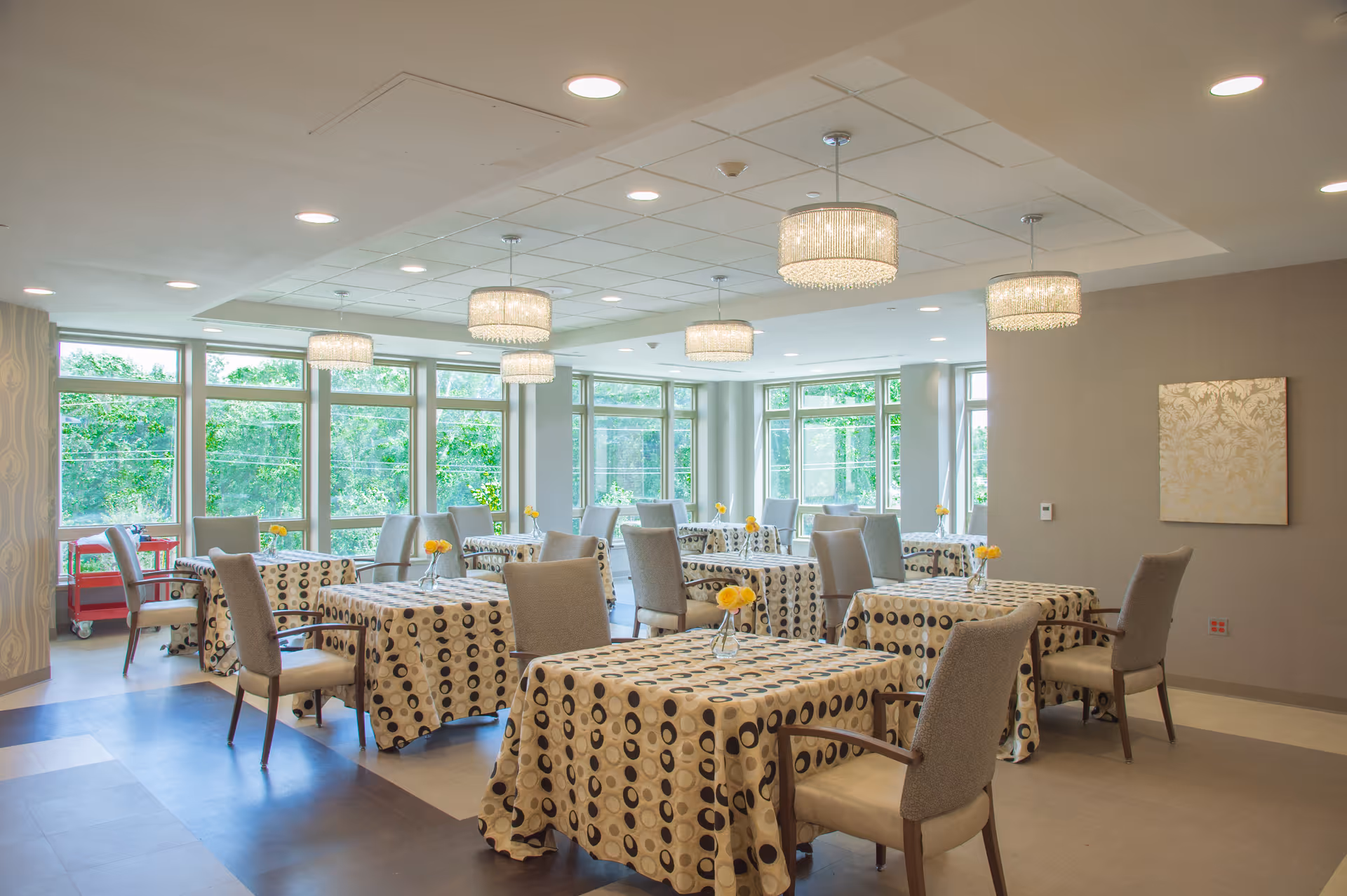 A bright and spacious dining room with multiple tables covered in patterned tablecloths. Each table has a small vase with yellow flowers. Comfortable upholstered chairs surround the tables. Large windows let in natural light and show greenery outside. Modern chandeliers hang from the ceiling, and a piece of artwork is displayed on one wall.